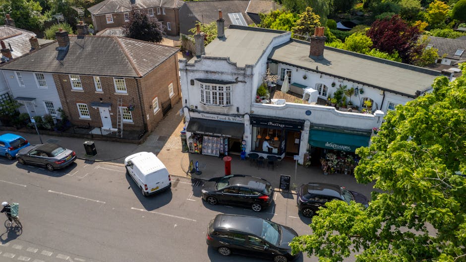 An aerial view of Hornchurch High Street showing a row of small shops with white and brick facades. In front, there are several parked cars along the street, including a white van, a black sedan, and