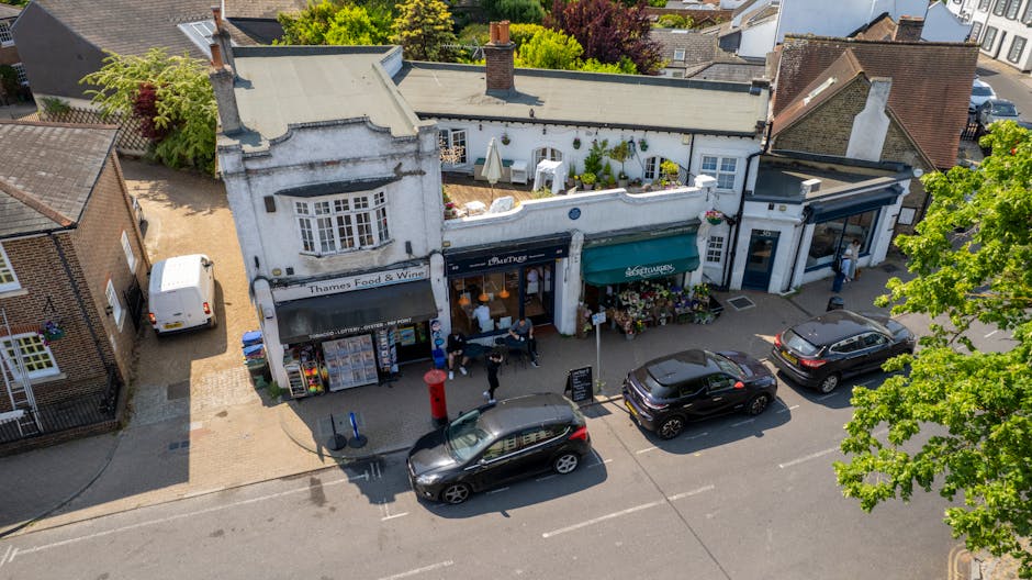 An aerial view of Hornchurch High Street showing a row of small shops with white and brick facades. In front, there are several parked cars along the street, including a white van, a black sedan, and