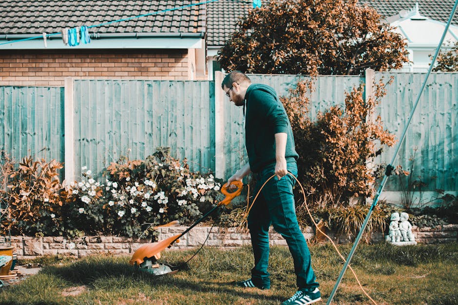 A man with short brown hair and glasses is engaged in garden maintenance in a backyard, using a cordless electric grass trimmer to trim the edge of a lush, green lawn. He is dressed in a dark hoodie a